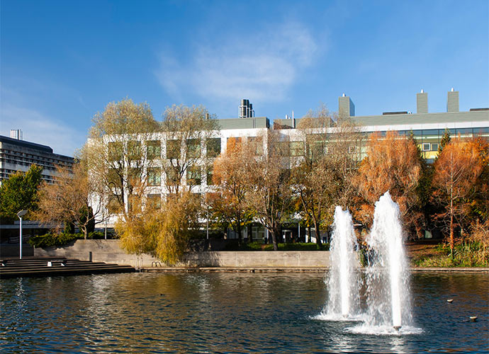 University College Dublin Science building behind a lake with two water fountains. Trees with autumn-coloured leaves line the water’s edge, and the sky is clear and blue.